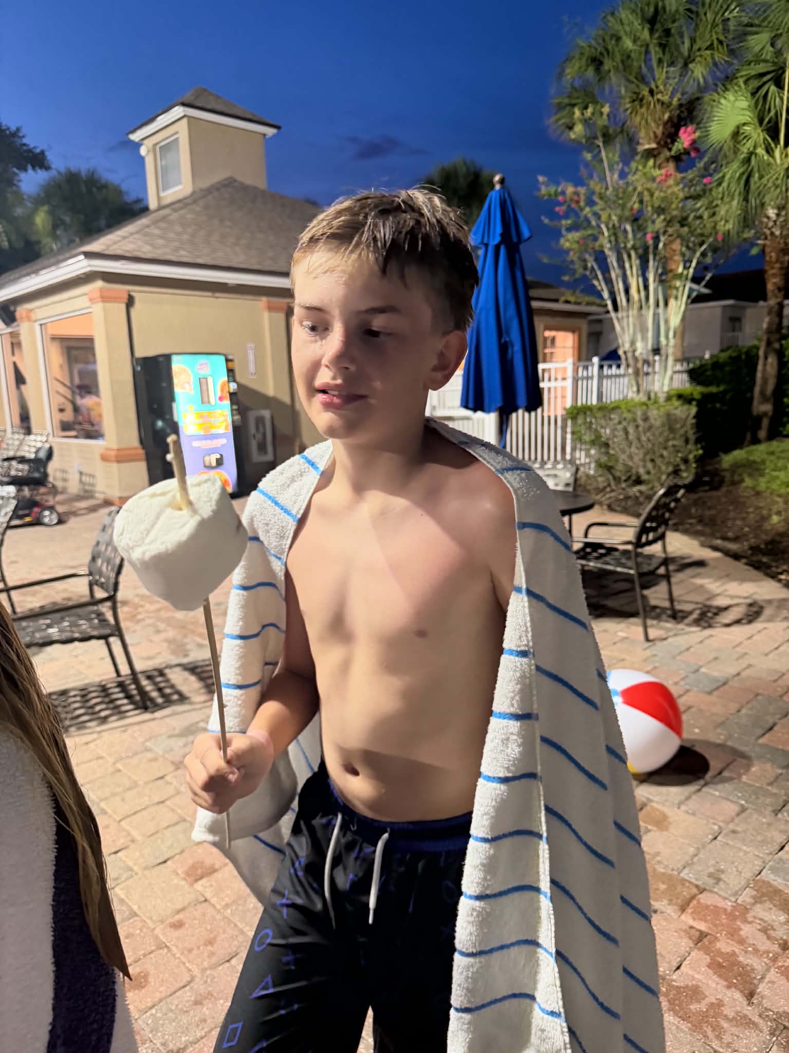 A shirtless boy with wet hair wrapped in a blue-striped towel holds a giant marshmallow on a stick on the resort pool deck at dusk, with resort buildings, a vending machine, and palm trees behind him