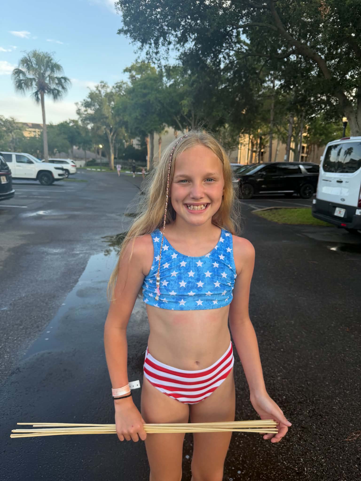 A smiling girl in a patriotic red, white, and blue swimsuit holds several long roasting sticks in a wet resort parking area with parked cars and palm trees behind her