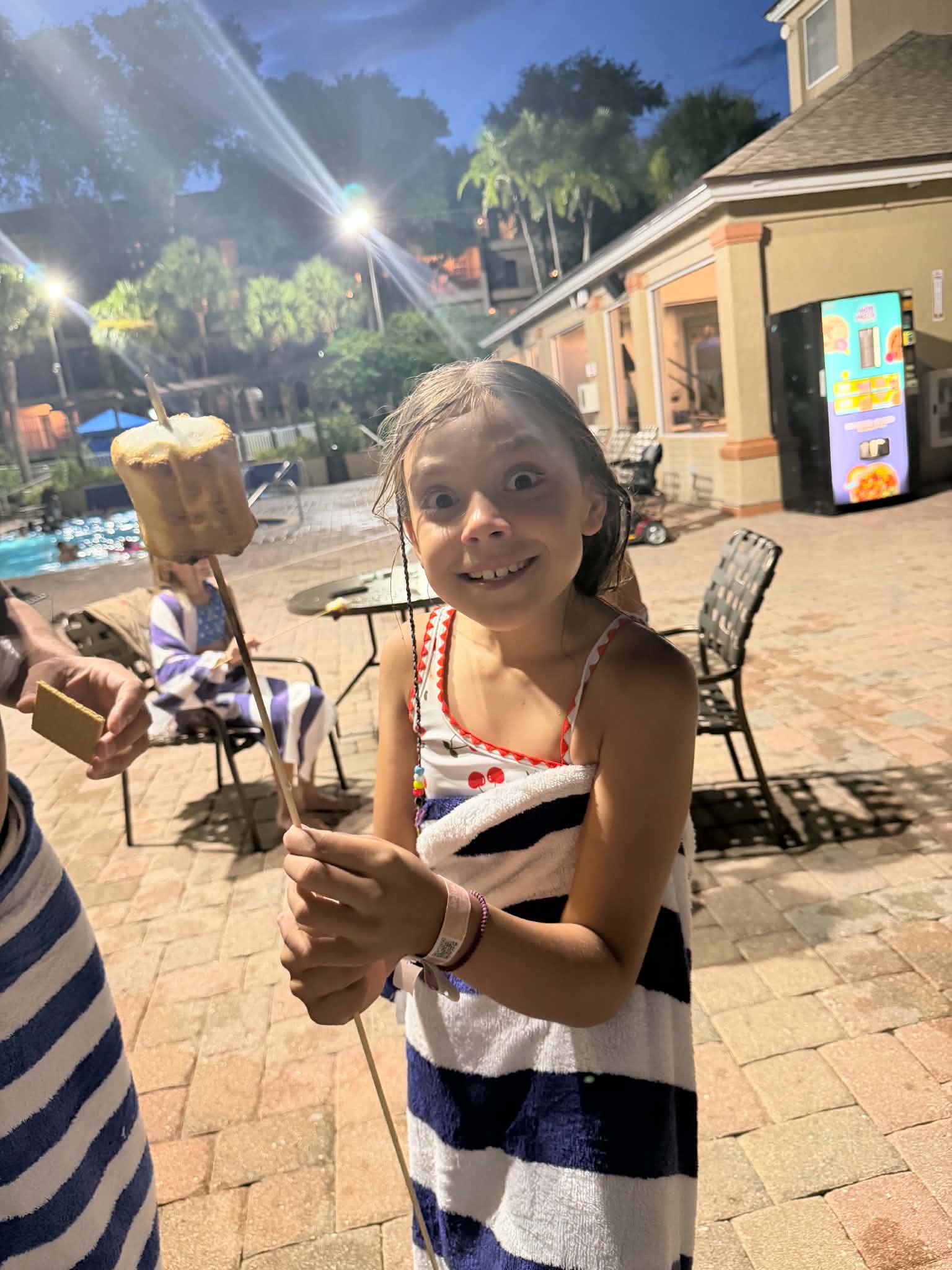 A girl holding a toasted marshmallow on a skewer stands beside the resort pool at night