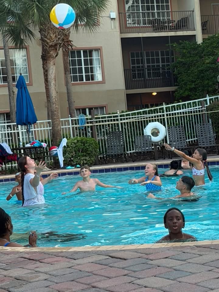 Children and adults play with a large colorful beach ball in the resort pool in front of palm trees and hotel balconies