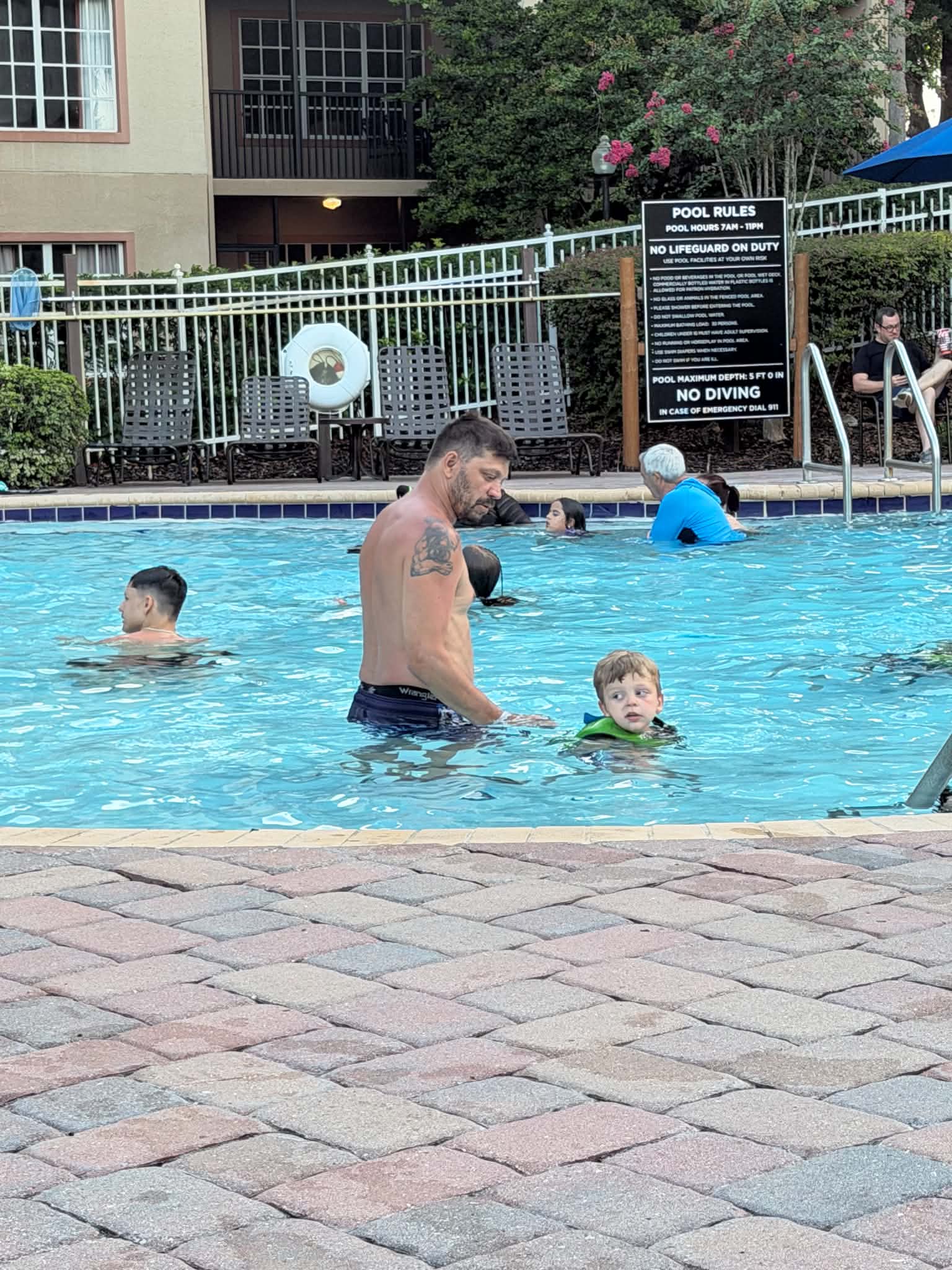 An adult stands in the resort pool beside a young child in a green flotation vest, with a pool rules sign and lounge chairs behind them