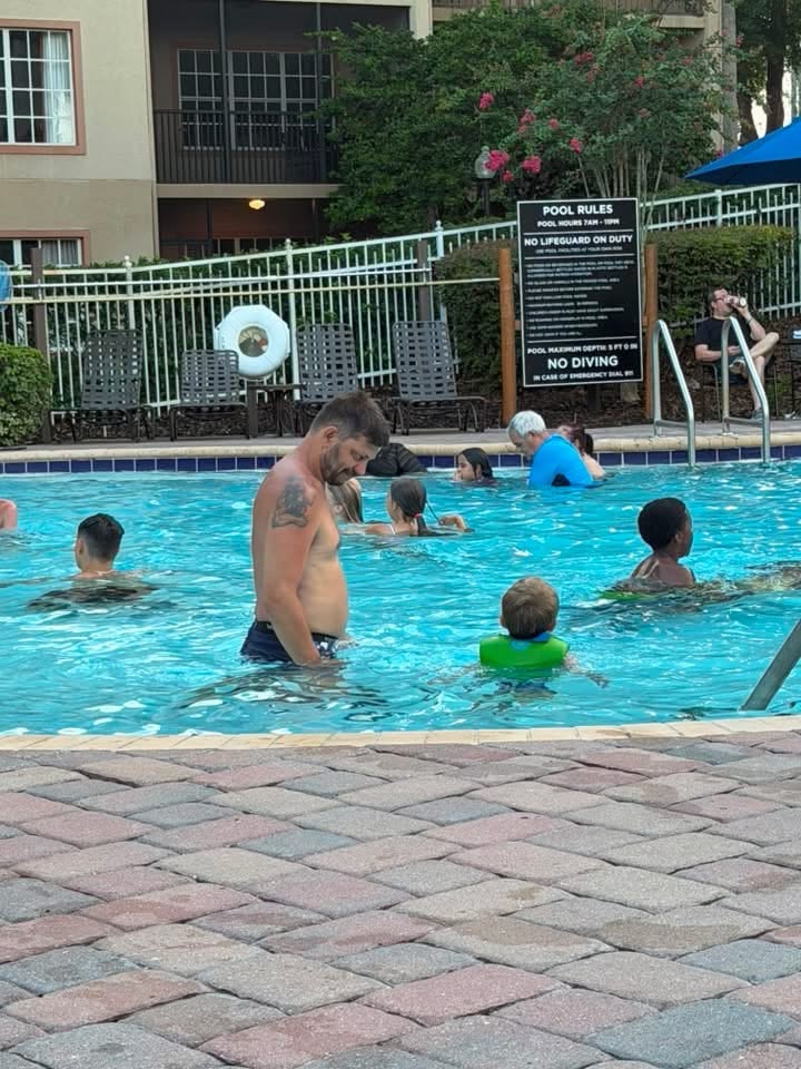 Families swim in the resort pool near lounge chairs and a pool rules sign