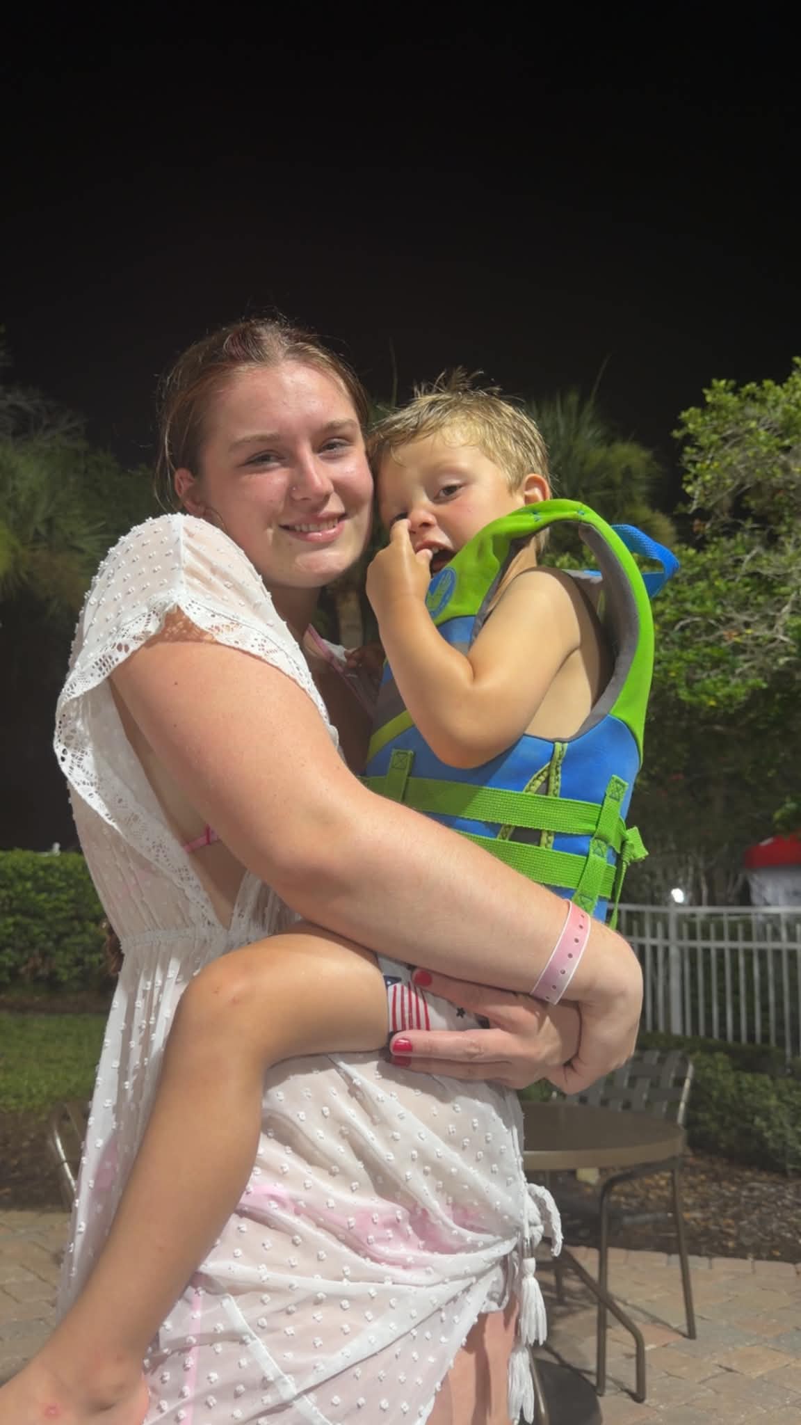 A young woman in a white cover-up holds a child in a green and blue flotation vest near the resort pool at night, with tropical landscaping and patio furniture behind them