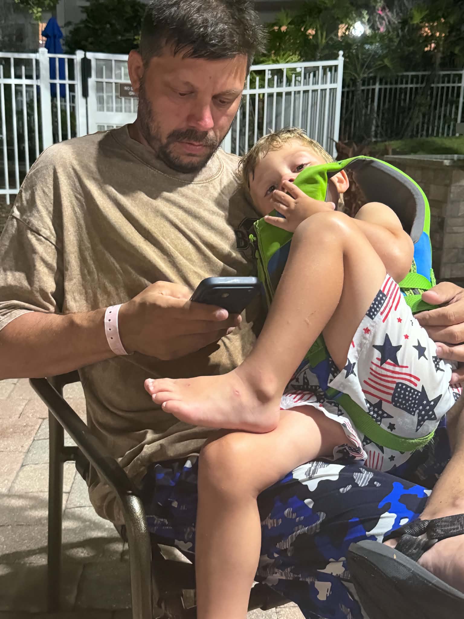 An adult holds a tired child in a green flotation vest beside the resort pool at night