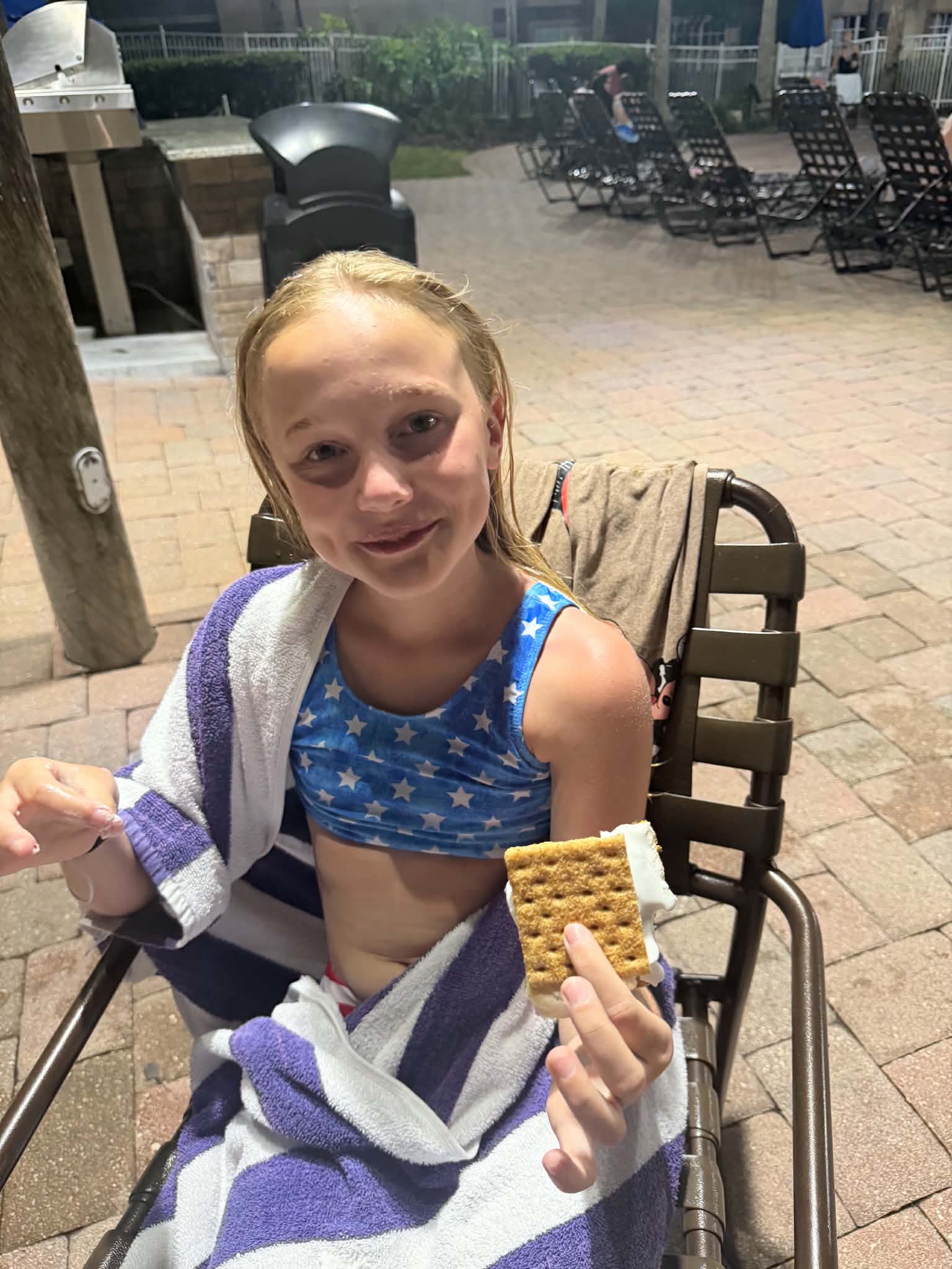 A girl wrapped in a striped towel holds a s'more while sitting on a patio chair beside the resort pool at night