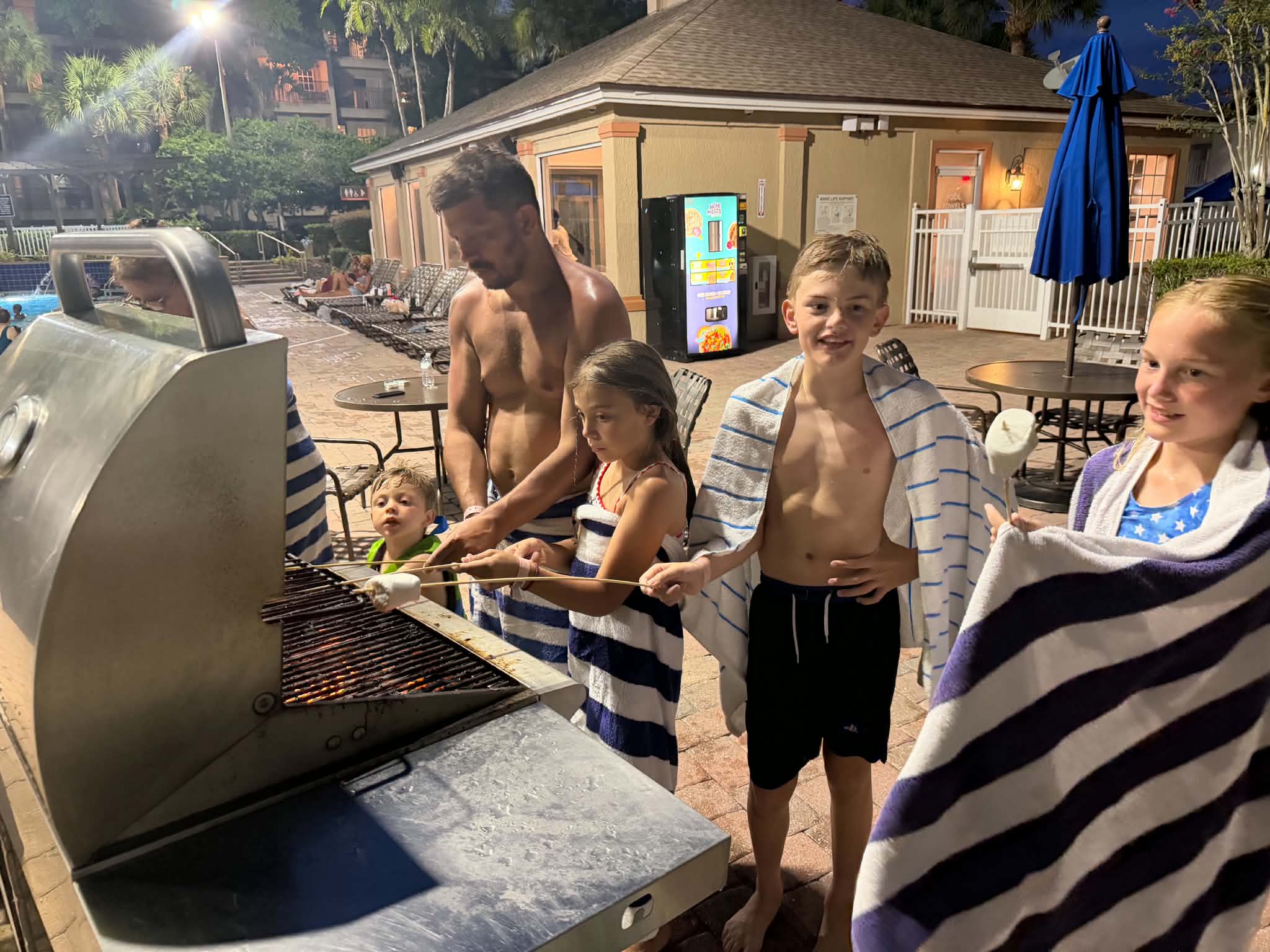 A man and several children gathered around the poolside grill at night roasting marshmallows on long skewers