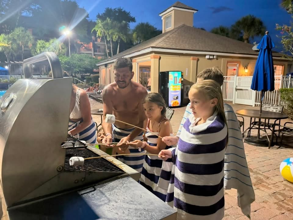 Kids wrapped in pool towels roast marshmallows on skewers beside the resort pool at night