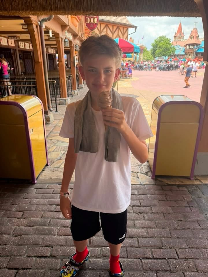 A boy stands in a shaded Disney park area eating an ice cream treat, with Fantasyland-style buildings and crowds in the background