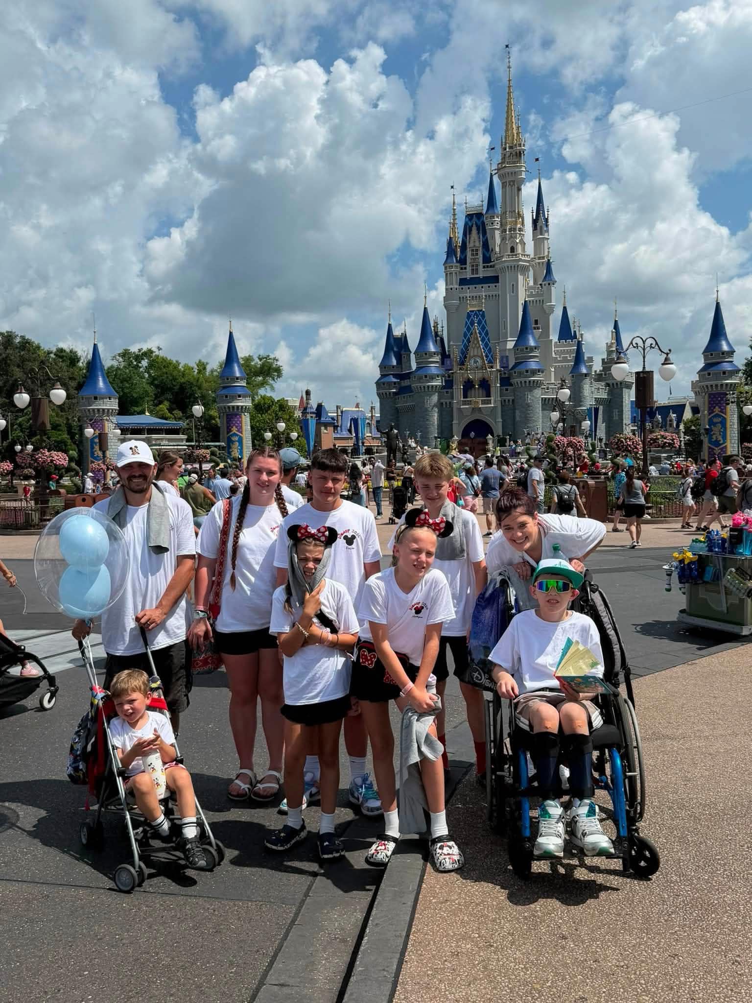 A family group in matching white Disney shirts poses in front of Cinderella Castle on a sunny day, with a blue Mickey balloon, stroller, and wheelchair in the foreground