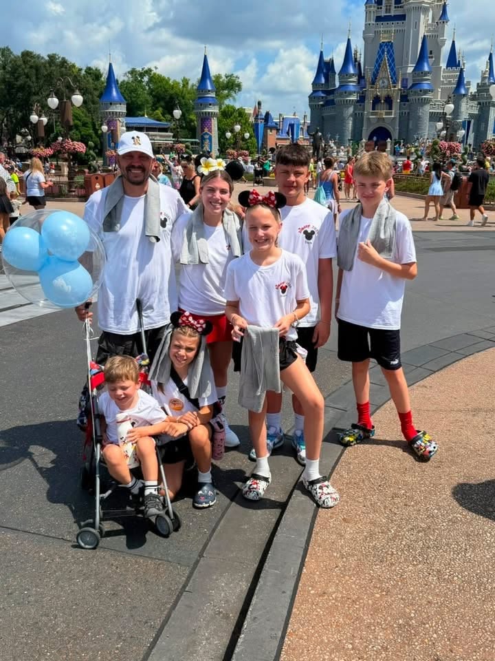 A smaller family group in matching white Disney shirts poses near Cinderella Castle on a sunny day, with a blue Mickey balloon and stroller beside them
