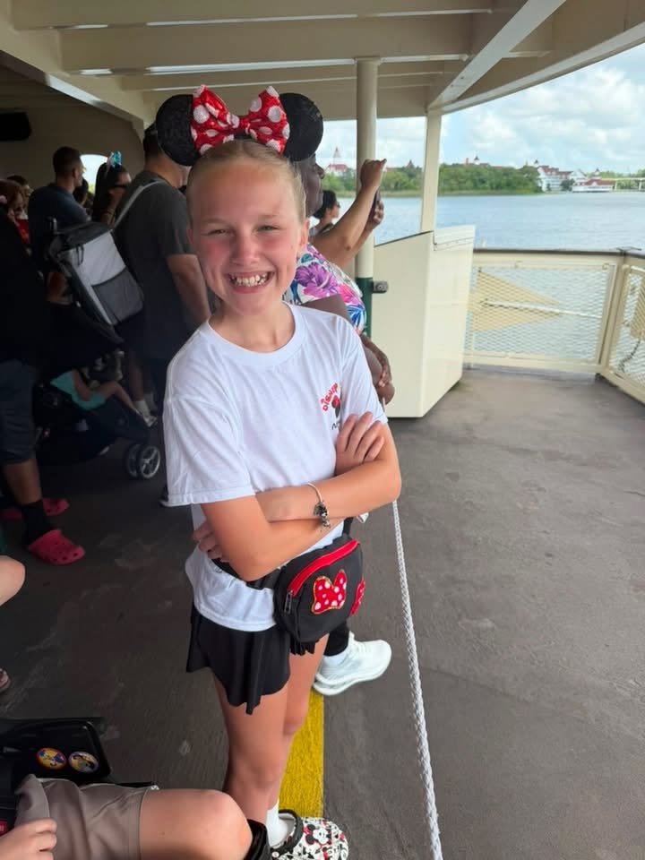 A smiling girl wearing Minnie ears stands on a Disney ferry deck with water and resort buildings in the background