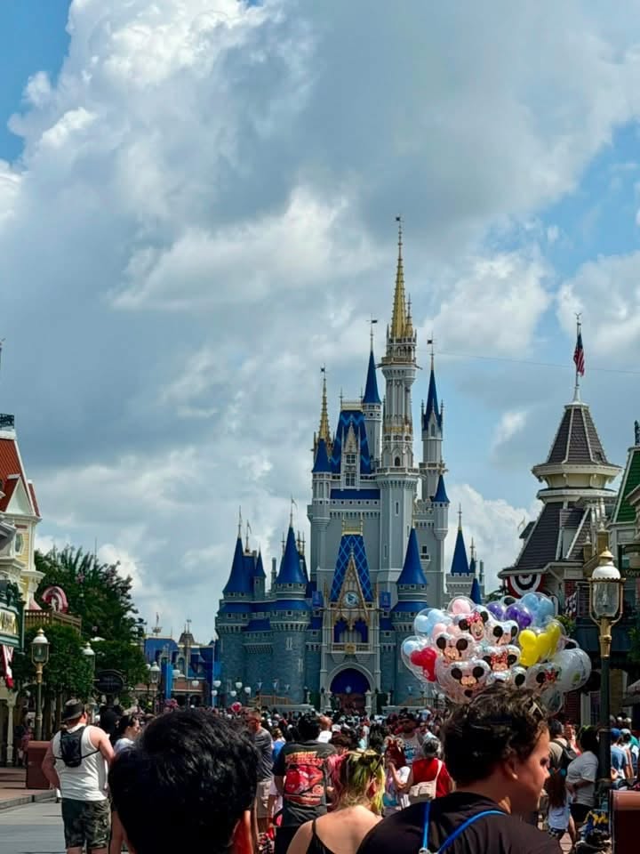 Crowds walk down Main Street U.S.A. toward Cinderella Castle under a cloudy blue sky, with colorful Mickey balloons visible on the right