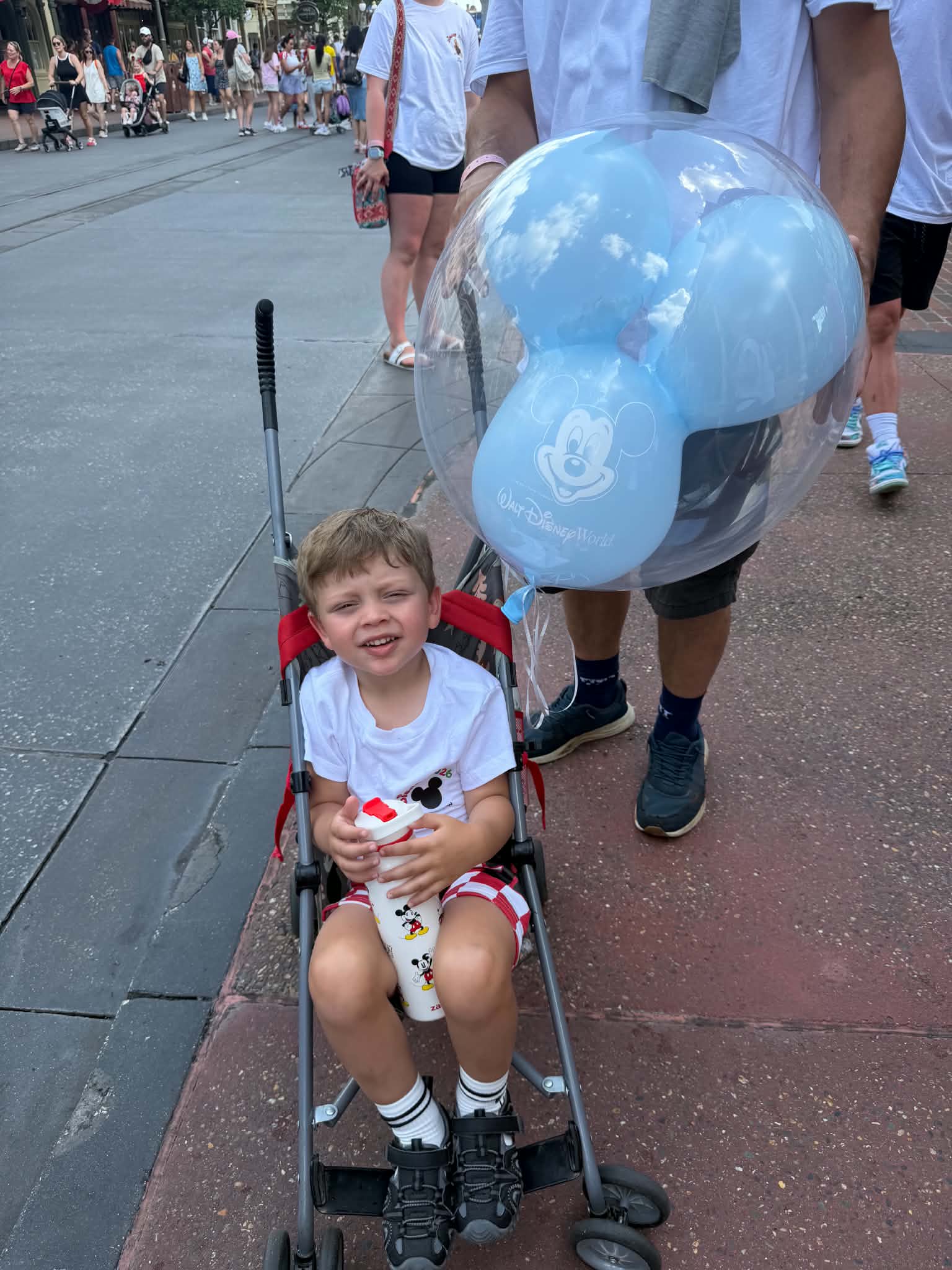 Young boy in a stroller holding a Mickey cup with a blue Mickey balloon beside him on Main Street