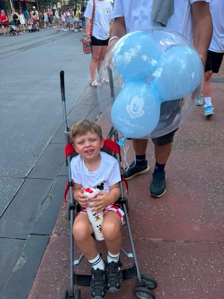 A young child sits in a stroller at Magic Kingdom holding a Mickey cup, with a clear blue Mickey balloon floating beside them